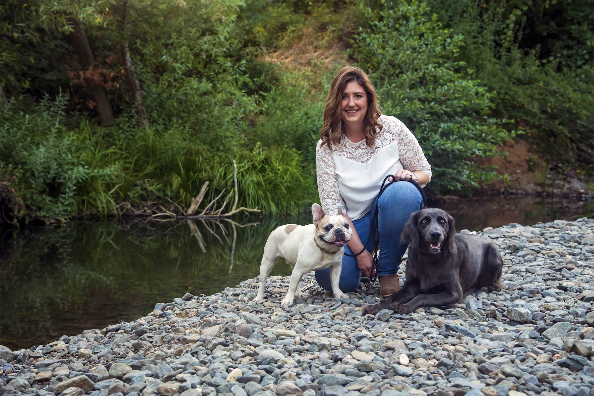 Trisha Schaeffer smiling and kneeling on a river's rocky shore, holding two dogs on leash.